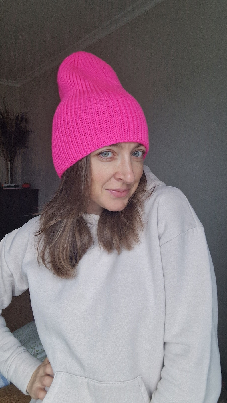 Op de afbeelding: Een felroze, geribbelde gebreide beanie. De hoed heeft een effen kleur en een losse pasvorm. De hoed wordt gedragen met een witte hoodie.