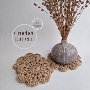 May include: Two crocheted beige coasters with a flower design. A vase with dried lavender is sitting on top of one of the coasters.