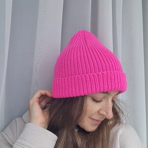 Op de afbeelding: Een felroze gebreide beanie-muts. De muts is gemaakt van een geribbelde gebreide stof en heeft een klassiek, nauwsluitend ontwerp. De heldere kleur maakt het een gedurfd modeaccessoire. De muts wordt in een close-up getoond.
