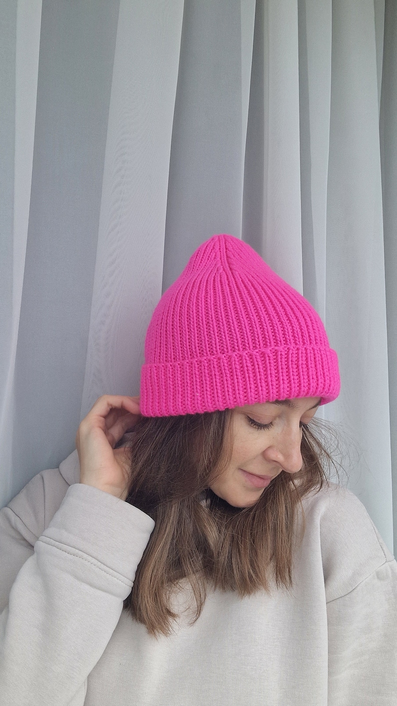 Op de afbeelding: Een felroze gebreide beanie-muts. De muts is gemaakt van een geribbelde gebreide stof en heeft een klassiek, nauwsluitend ontwerp. De heldere kleur maakt het een gedurfd modeaccessoire. De muts wordt in een close-up getoond.