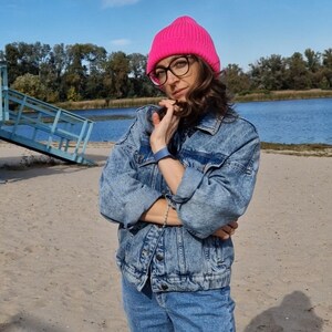 Op de afbeelding: Een persoon draagt een felroze gebreide beanie, een bril met zwart montuur en een blauwe spijkerjas en jeans. De achtergrond toont een zandstrand, een blauw meer en een blauwe lucht met bomen.
