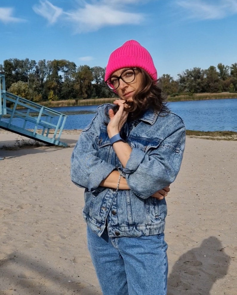 Op de afbeelding: Een persoon draagt een felroze gebreide beanie, een bril met zwart montuur en een blauwe spijkerjas en jeans. De achtergrond toont een zandstrand, een blauw meer en een blauwe lucht met bomen.