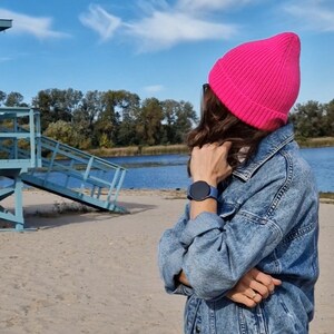 Op de afbeelding: Een persoon draagt een felroze gebreide beanie en een blauw spijkerjack. Een blauwe smartwatch is zichtbaar om de pols. De achtergrond omvat een zandstrand, een blauwe strandwachtpost en een watermassa onder een blauwe lucht.