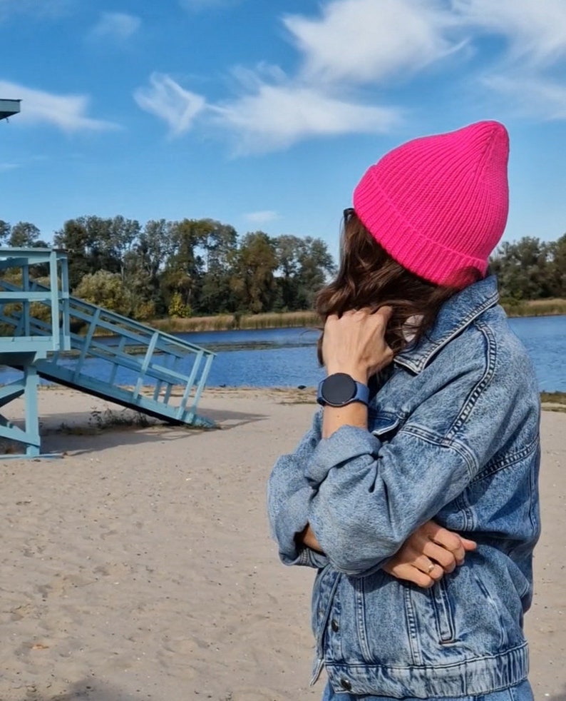 Op de afbeelding: Een persoon draagt een felroze gebreide beanie en een blauw spijkerjack. Een blauwe smartwatch is zichtbaar om de pols. De achtergrond omvat een zandstrand, een blauwe strandwachtpost en een watermassa onder een blauwe lucht.