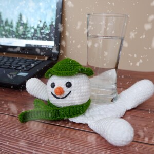 Könnte beinhalten: Ein weißer Häkel-Schneemann mit grünem Hut und Schal, der auf einem Holztisch mit einem Glas Wasser und einem Laptop im Hintergrund sitzt.