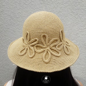 Può includere: Un cappello da sole in crochet beige con un'ampia tesa e tre dettagli a forma di fiore attorno alla corona. Il cappello è realizzato con un punto testurizzato e ha un design semplice ed elegante.