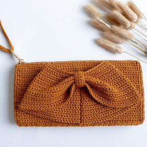 May include: A brown crocheted clutch bag with a large bow on the front and a long strap.