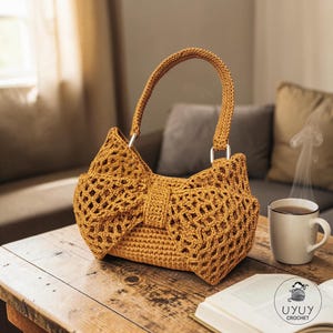 May include: A mustard-coloured crochet handbag with a bow-shaped design. The bag has a structured shape with a handle and silver-coloured rings. The open-weave pattern is visible. The bag is sitting on a wooden table.