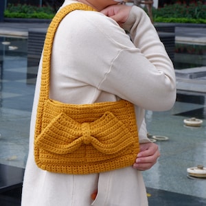 May include: A mustard yellow crocheted handbag with a large bow detail. The bag is worn over the shoulder of a person.