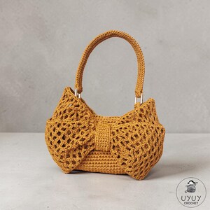 Può includere: Borsa a mano all'uncinetto giallo senape con design a forma di fiocco. La borsa presenta una forma strutturata con manico e finiture argentate. L'uncinetto ha un motivo aperto e traforato. La borsa è appoggiata su una superficie neutra.