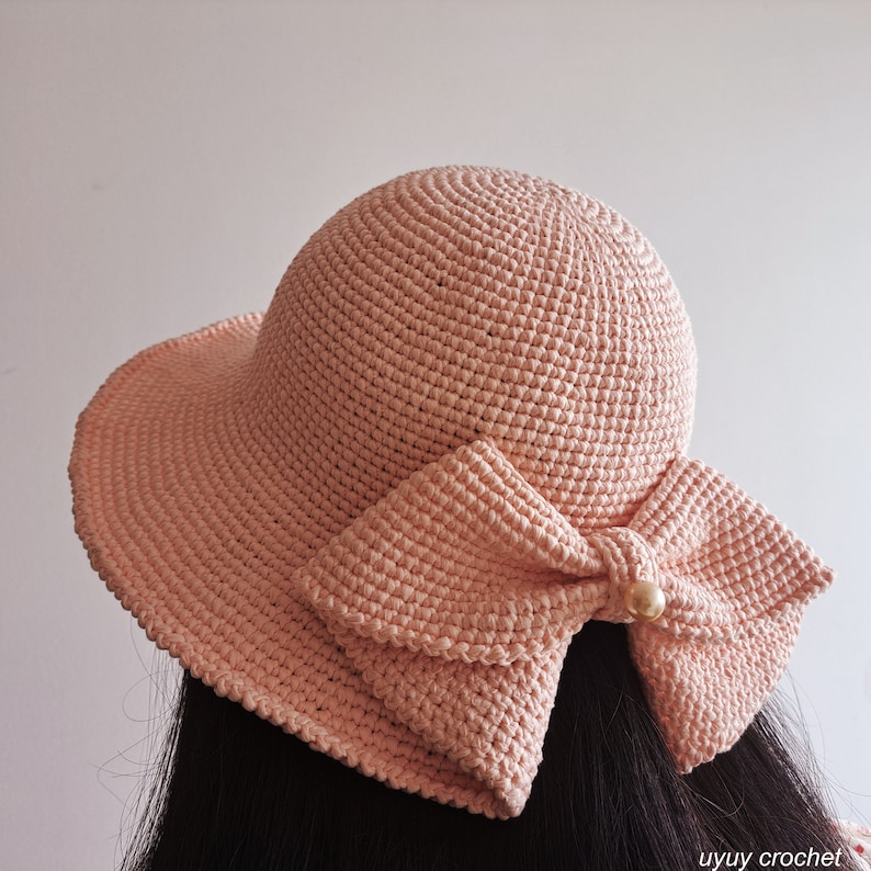 May include: A pink crocheted sun hat with a large bow on the back. The bow is also crocheted and has a pearl embellishment in the center.