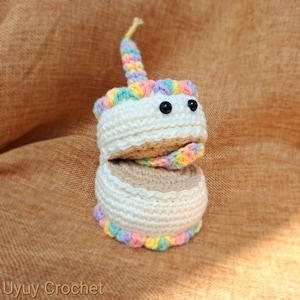 以下が含まれることがあります： 風変わりなデザインの、かぎ針編みのケーキ型おもちゃ。白いケーキは、上部と下部に虹色のトリムがあり、虹色の舌と黄色のろうそくが付いています。黒いボタンの目が遊び心のある外観を与えています。