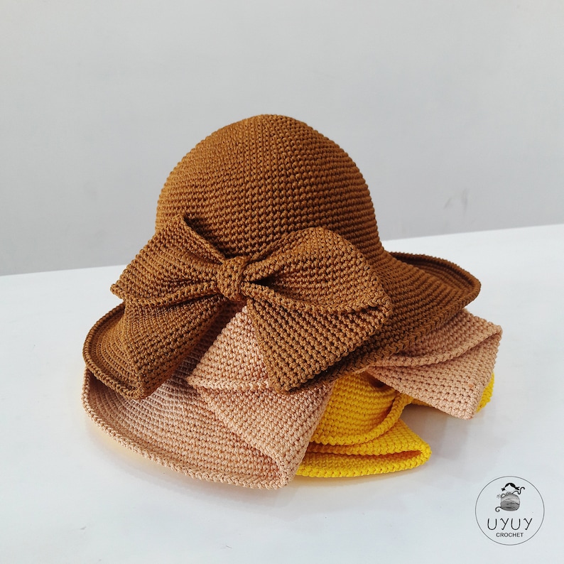 May include: A brown crocheted sun hat with a large brown bow. The hat is layered with a beige crocheted hat and a yellow crocheted hat.