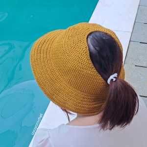 May include: A crocheted sun hat in a mustard yellow color with a large brim and an open back. The hat is worn by a person with long brown hair.