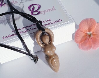 May include: A brown wood carved goddess pendant on a black cord necklace. The pendant is shaped like a woman with her arms raised above her head. The pendant is on a white surface with a pink flower in the background.