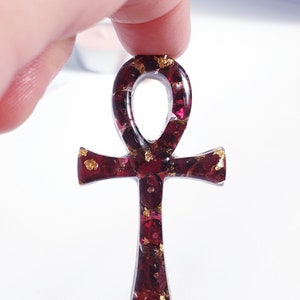 May include: A red and gold Ankh pendant made with resin and embedded with small red gemstones. The pendant is held in a hand.