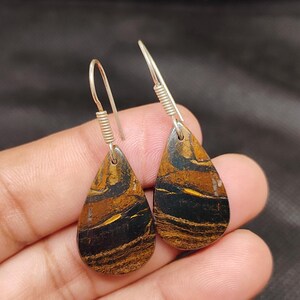 Può includere: Un paio di orecchini a goccia dall'aspetto di pietra naturale. Gli orecchini presentano ganci argentati e un motivo a bande nere e dorate. I gioielli sono esposti su uno sfondo scuro.