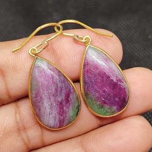 Peut inclure: Boucles d'oreilles pendantes en forme de goutte d'eau dorées, ornées de pierres précieuses de rubis naturelles. Les rubis présentent des teintes roses, violettes et vertes. Les boucles sont suspendues à des crochets dorés.