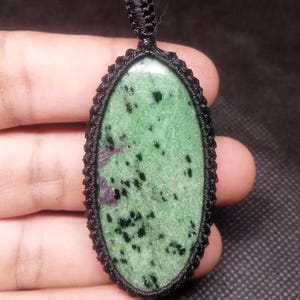 May include: Oval pendant with a green stone, black and red inclusions, and a black macrame setting. The pendant is suspended from a woven loop. The design showcases a natural gemstone.