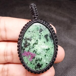 May include: Oval-shaped pendant featuring a green stone with black and pink inclusions, encased in a black macrame setting. The pendant has a loop at the top for hanging. The stone's surface has a speckled appearance.