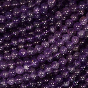 May include: A collection of smooth, round amethyst beads arranged in multiple parallel strands. The beads exhibit varying shades of deep purple and violet, featuring natural internal inclusions and a polished, reflective surface finish.