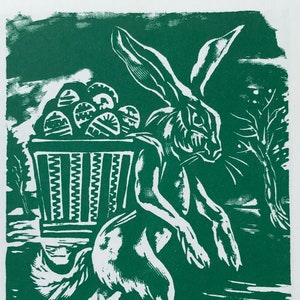 Pode incluir: Uma gravura em linóleo verde e branco retrata um coelho carregando uma cesta de ovos de Páscoa decorados. O coelho está andando para a direita, com uma cesta nas costas. O fundo inclui árvores e um céu nublado.