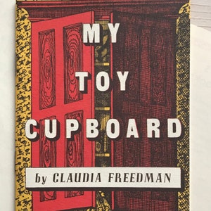 May include: A book cover with the title "MY TOY CUPBOARD" in large white letters. The cover features a red door with ajar, revealing a dark brown interior. The author's name, "CLAUDIA FREEDMAN," is printed below the title.