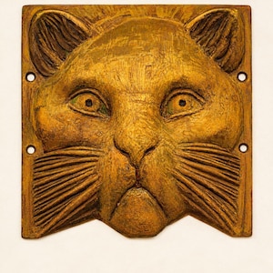 Könnte beinhalten: Eine quadratische, goldbraune Wandplatte mit einem Relief eines Katzengesichts. Die katzenartigen Merkmale umfassen spitze Ohren, große Augen und detaillierte Schnurrhaare. In jeder Ecke des quadratischen Rahmens befindet sich ein kleines Loch.