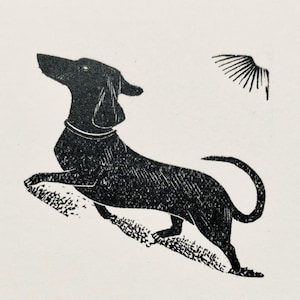 Puede incluir: Un grabado en linóleo en blanco y negro de un perro saltando hacia arriba, con un pequeño gráfico de ala en la esquina superior derecha. El perro, con collar, mira hacia arriba, posiblemente persiguiendo el ala. La impresión tiene un aspecto texturizado.