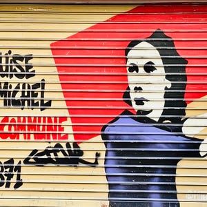 Może przedstawiać: Mural na beżowej metalowej żaluzji przedstawiający stylizowany portret osoby z ciemnymi włosami w fioletowej koszuli na czerwonym prostokątnym tle. Czarny napis głosi LOUISE MICHEL, LA COMMUNE i 1871, z dodatkowymi czarnymi graffiti.