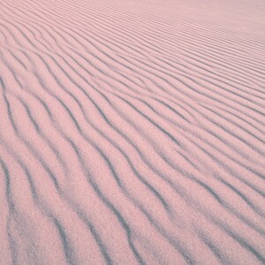May include: Close-up of sand dunes with a rippled texture. The sand is a soft pink color, with darker, wavy lines creating a pattern across the surface. The image captures the natural beauty of the desert landscape.