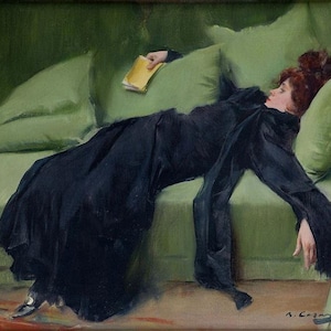 May include: A woman in a black dress lies on a green sofa with a yellow book in her hand. The sofa has cushions and the woman's hair is red.