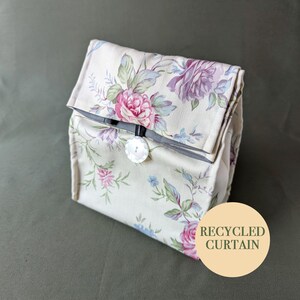 May include: A white fabric lunch bag with a floral pattern and a white button closure. The bag is made from recycled curtain fabric. The text "RECYCLED CURTAIN" is printed on a beige circle.
