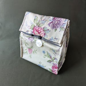 May include: A beige fabric lunch bag with a floral pattern featuring pink, purple, and blue roses. The bag has a black button closure.
