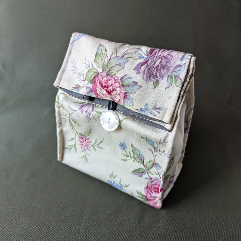May include: A beige fabric lunch bag with a floral pattern featuring pink, purple, and blue roses. The bag has a black button closure.