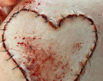 May include: A heart-shaped wound with black stitches and simulated blood splatters. The heart outline is stitched with black thread, and the surrounding skin has a pale tone. The image evokes a sense of horror or medical theme.