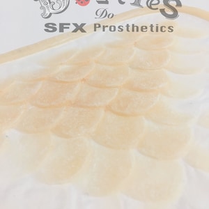 May include: A close-up of a prosthetic wing with a scale-like design. The wing is a light beige colour and is made of a flexible material. The text "Dotties Do SFX Prosthetics" is at the top of the image.