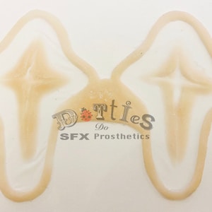 May include: A pair of prosthetic appliances with a light skin tone, featuring a central design and a curved outer edge. The text "Dotties Do SFX Prosthetics" is visible in the center.