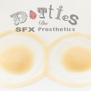 Puede incluir: Dos prótesis mamarias SFX de tono de piel claro, cada una rodeada por un borde fino y translúcido. La imagen incluye el texto "Dotties Do SFX Prosthetics" en una fuente decorativa, con un icono de mariquita.
