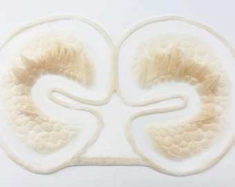 May include: A beige, symmetrical prosthetic appliance with a textured surface and a clear border. The design features two rounded sections connected by a central bridge, with a detailed, organic appearance. The object is set against a white background.