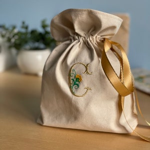 May include: A cream-coloured drawstring bag with a gold ribbon. The bag is embroidered with a gold letter "C" and a yellow flower.