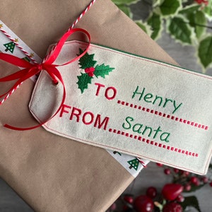May include: A Christmas gift wrapped in brown paper, adorned with a cream-coloured tag embroidered with "TO Henry FROM Santa" in green and red. The tag features a holly design. The gift is tied with a red ribbon and festive twine.