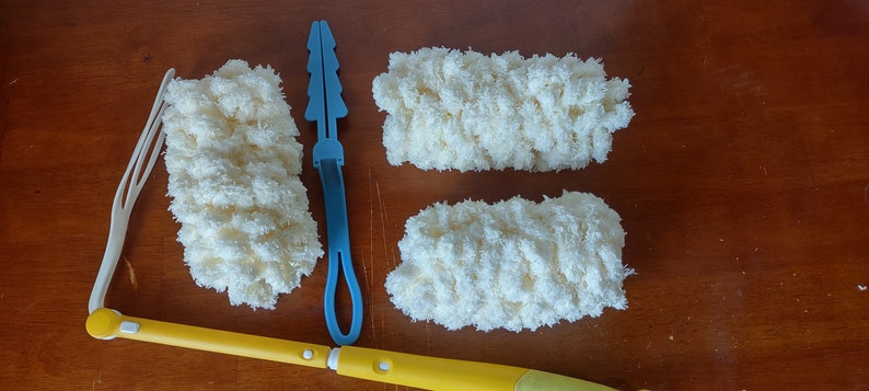 May include: A yellow and white duster with a long handle, a white plastic attachment, and three fluffy, cream-colored cleaning pads. A blue plastic tool is also included. The items are arranged on a wooden surface.