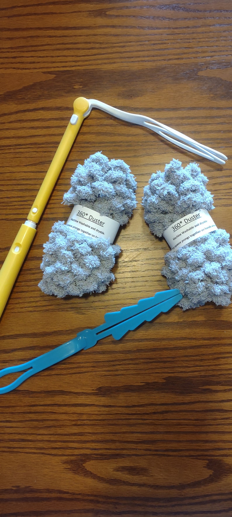 May include: Two gray microfiber dusters with white labels that read "360* Duster Machine Washable and dryable.  Secure prongs together on handle."  The dusters are on a wooden surface with a yellow and white duster handle and a blue plastic handle.