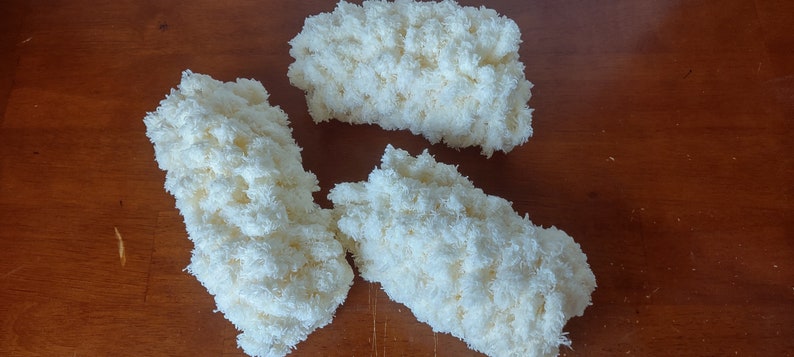 May include: Three fluffy, off-white, rectangular yarn pieces are arranged on a wooden surface. The yarn appears soft and textured, with a dense, cloud-like appearance. The pieces are likely for crafting or textile projects.