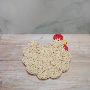 Puede incluir: Un posavasos de pollo tejido a crochet en color beige con detalles en rojo y amarillo.