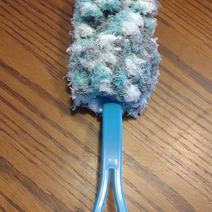 May include: A cleaning tool featuring a light blue plastic handle attached to a textured, scrubby head. The head is composed of yarn in a variegated color palette of white, grey, and teal, creating a dense, looped, and multi-colored surface texture.