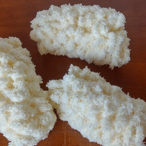 May include: Three fluffy, off-white yarn pieces arranged on a wooden surface. The yarn appears to be a soft, textured material, possibly for crafting or home decor. The pieces are irregularly shaped, suggesting a handmade or natural aesthetic.