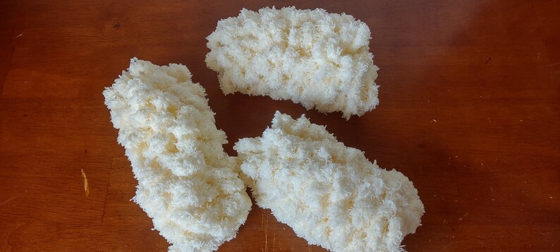 May include: Three fluffy, off-white yarn pieces arranged on a wooden surface. The yarn appears to be a soft, textured material, possibly for crafting or home decor. The pieces are irregularly shaped, suggesting a handmade or natural aesthetic.