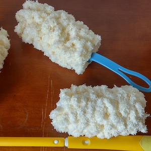 May include: A collection of cleaning dusters with fluffy, cream-colored microfiber heads. One duster has a yellow handle, another has a blue handle, and the third has a white handle. The dusters are arranged on a wooden surface.
